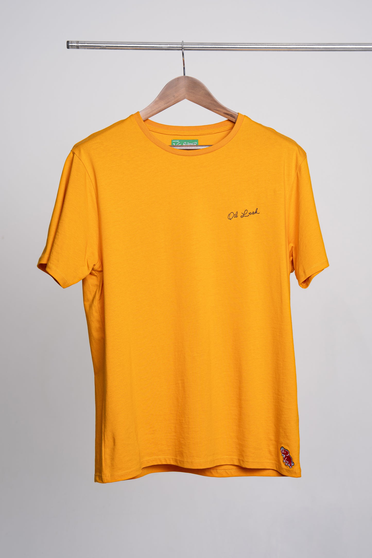 Yellow t-shirt with small embroidery that reads "oil leak" on a hanger against a gray background