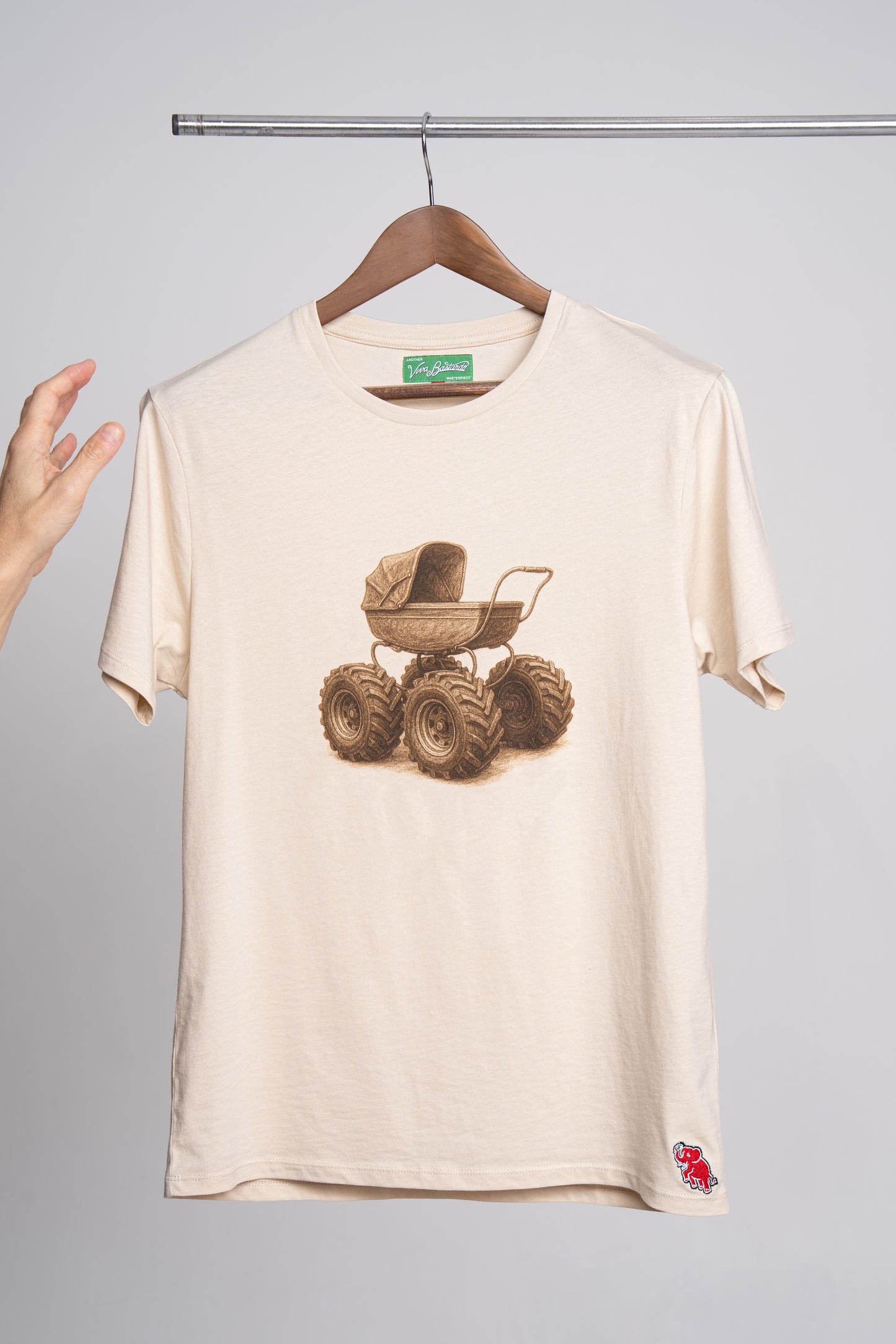 Beige t-shirt with a graphic of a baby carriage on tractor wheels, displayed on a hanger against a gray background.